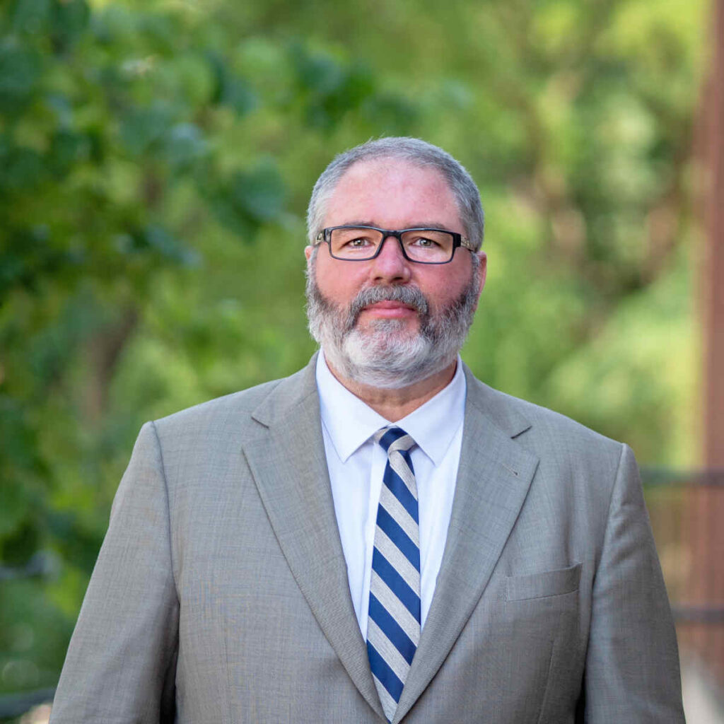 photo of man in a light grey suit wearing a blue diagonal striped tie. the man is wearing glasses and has a beard