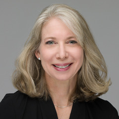 Bio photo of woman with shoulder length blond hair wearing a black jacket in front of a grey background.